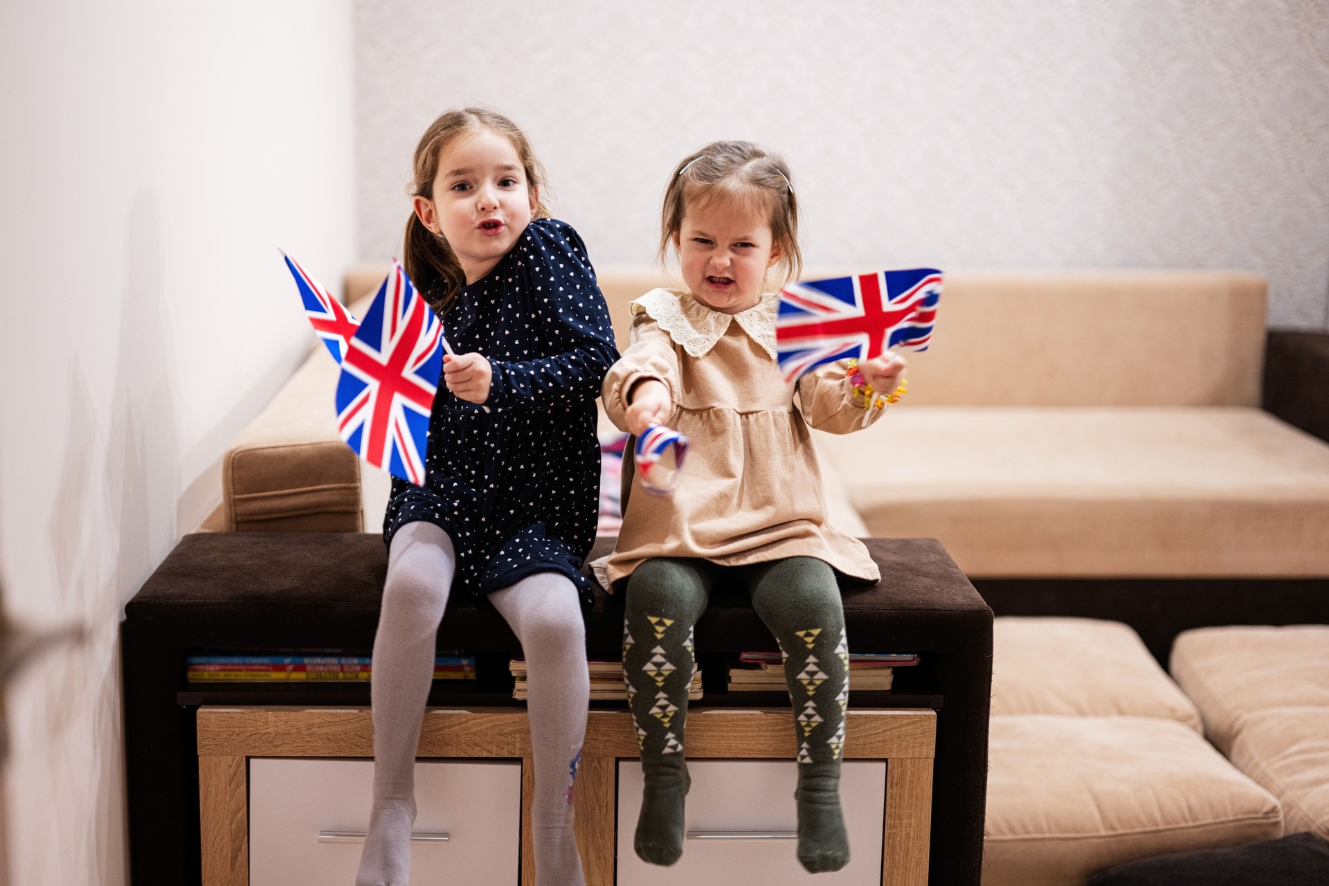 Two sisters are sitting on a couch at home with british flags on hands. Great Britain children girls with flag .